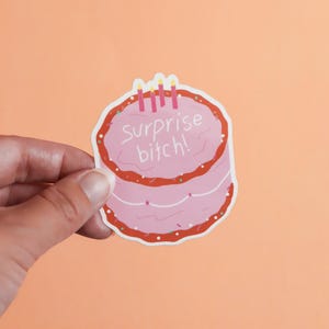 May include: A pink sticker in the shape of a cake with red frosting and three candles. The text "surprise bitch!" is written on the cake in white letters. The sticker is held by a hand against an orange background.