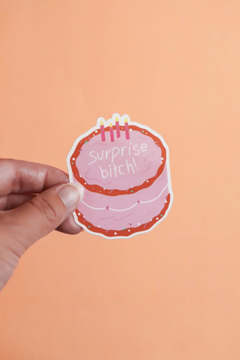 May include: A pink sticker in the shape of a cake with red frosting and three candles. The text "surprise bitch!" is written on the cake in white letters. The sticker is held by a hand against an orange background.