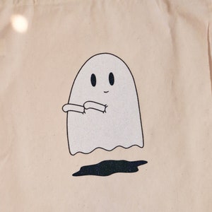 May include: A white canvas tote bag with a glittery ghost design. The ghost is smiling and has its arms crossed. The ghost is floating above a black glittery puddle.