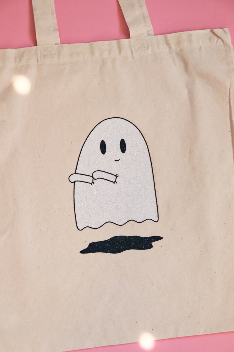 May include: A white canvas tote bag with a glittery ghost design. The ghost is smiling and has its arms crossed. The ghost is floating above a black glittery puddle.
