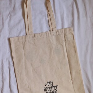 May include: A natural-colored tote bag with black handles and the text "..day dreamer studios.." printed on the front.