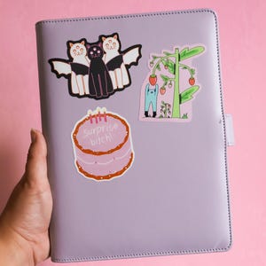 May include: A lilac-colored planner adorned with stickers. The stickers include a cake with the text "Surprise bitch!", a trio of cats with bat wings, and a cartoon of a person tending a strawberry plant. The background is a solid pink.