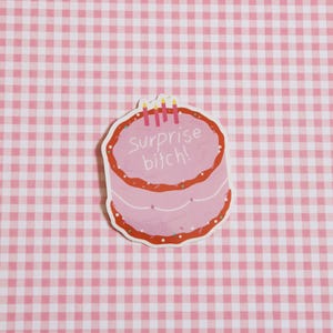 May include: A pink sticker in the shape of a cake with red frosting and sprinkles. The text "Surprise bitch!" is written on the cake. Three lit candles are on top of the cake. The background is a pink and white checkered pattern.