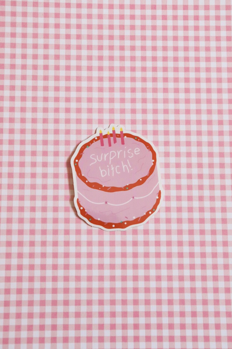 May include: A pink sticker in the shape of a cake with red frosting and sprinkles. The text "Surprise bitch!" is written on the cake. Three lit candles are on top of the cake. The background is a pink and white checkered pattern.