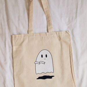 May include: A beige canvas tote bag with a white ghost design. The ghost has two eyes and a small smile. The ghost is holding its arms out in front of it.