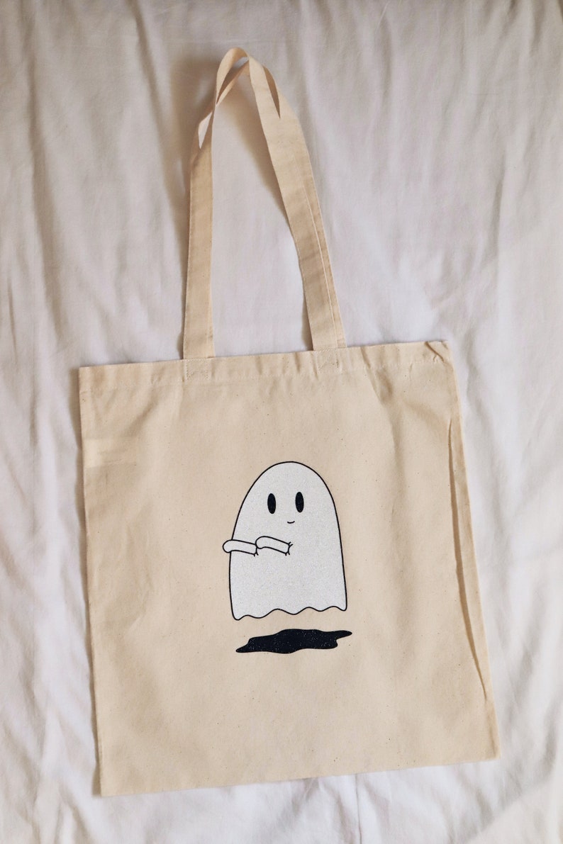 May include: A beige canvas tote bag with a white ghost design. The ghost has two eyes and a small smile. The ghost is holding its arms out in front of it.