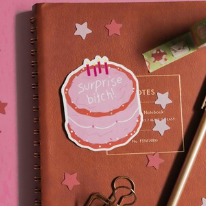 May include: A pink cake sticker with the text "Surprise bitch!" and three candles is placed on a brown notebook. The notebook is surrounded by pink star-shaped confetti, a gold binder clip, and a pen. A roll of decorative tape is also visible.