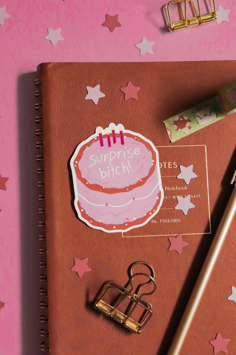 May include: A pink cake sticker with the text "Surprise bitch!" and three candles is placed on a brown notebook. The notebook is surrounded by pink star-shaped confetti, a gold binder clip, and a pen. A roll of decorative tape is also visible.