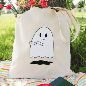 May include: A white canvas tote bag with a black and white illustration of a ghost with arms outstretched. The bag is sitting on a striped picnic blanket with a book and sunglasses.