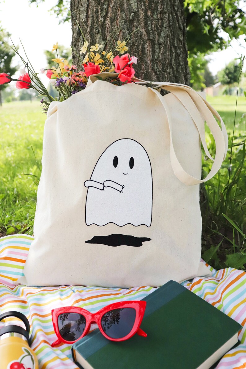 May include: A white canvas tote bag with a black and white illustration of a ghost with arms outstretched. The bag is sitting on a striped picnic blanket with a book and sunglasses.