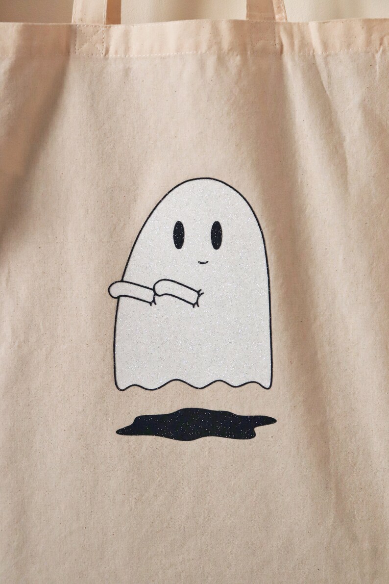 May include: A white tote bag with a glittery ghost design. The ghost is smiling and has its arms outstretched.