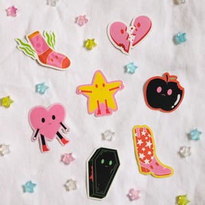 May include: A set of eight colorful stickers featuring various cartoon characters, including a sock, a heart, a star, a boot, a coffin, and an apple. The stickers are on a white background with scattered pink, blue, and yellow stars.