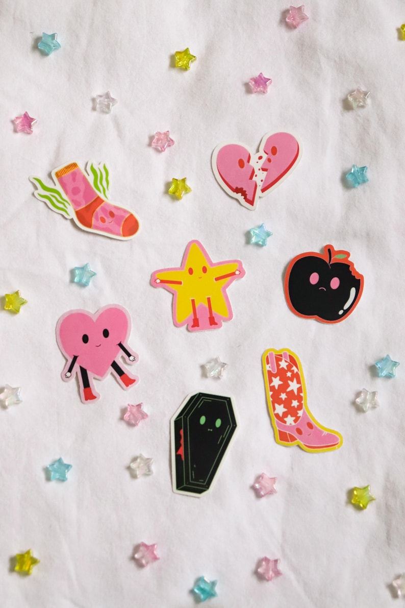 May include: A set of eight colorful stickers featuring various cartoon characters, including a sock, a heart, a star, a boot, a coffin, and an apple. The stickers are on a white background with scattered pink, blue, and yellow stars.