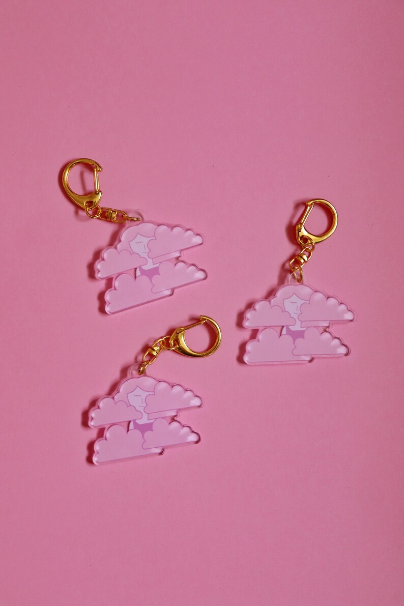 May include: Three pink and white cloud-shaped keychains with a gold chain and ring. Each keychain features a small cartoon person inside the clouds.