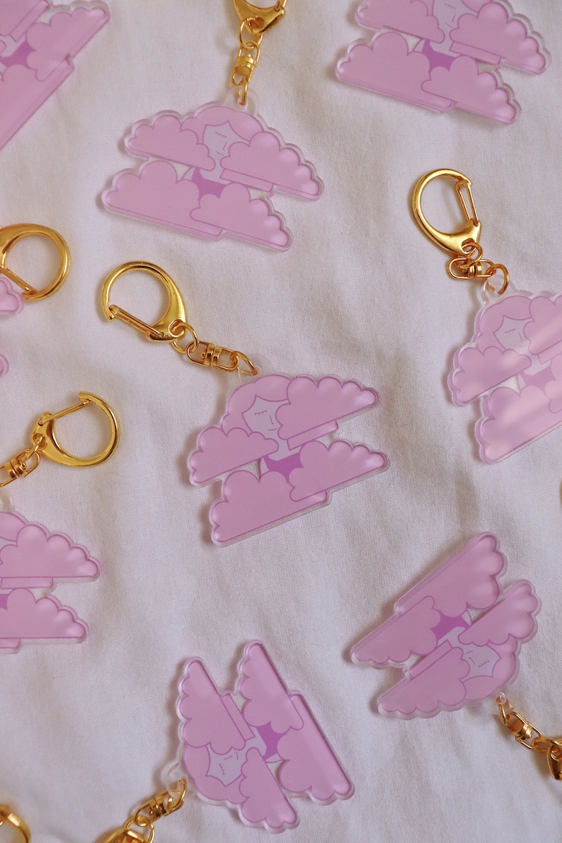 May include: A set of pink and gold keychains featuring a cartoon character in clouds. The keychains are made of clear acrylic and have a gold metal ring.