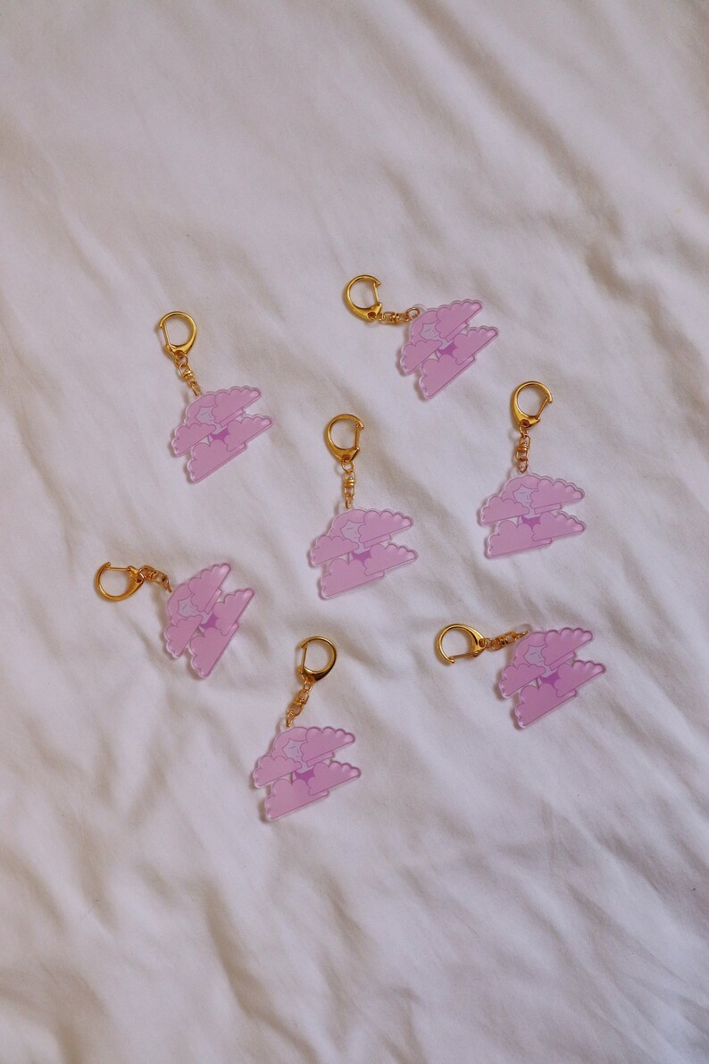 May include: Seven pink and purple cloud-shaped keychains with gold rings. Each keychain features a double-layered cloud with a star in the center.
