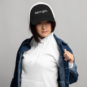 May include: Black baseball cap with the text "barn girl." The cap has a curved brim and a distressed look. The hat is worn with a white hoodie and a denim jacket.