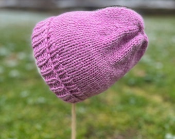 May include: A pink knitted beanie hat displayed on a wooden stand. The hat is made of a soft, textured yarn and has a simple, classic design.
