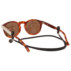 Puede incluir: Gafas de sol redondas de carey con lentes marrones y cordón negro. Las gafas tienen un acento plateado en la patilla y un pequeño adorno decorativo. El cordón está sujeto a las patillas con presillas de goma negra.