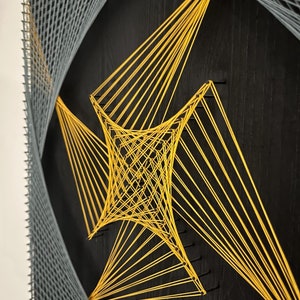 May include: A black wooden board with a geometric design created with string. The design features a large, yellow diamond shape in the center, surrounded by a smaller, gray diamond shape.