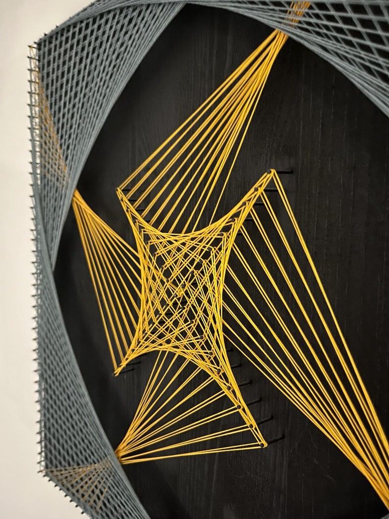 May include: A black wooden board with a geometric design created with string. The design features a large, yellow diamond shape in the center, surrounded by a smaller, gray diamond shape.