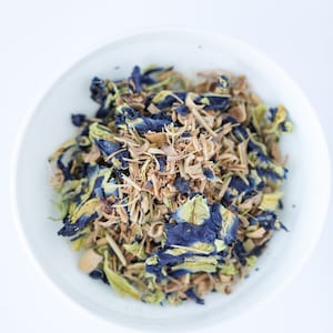 May include: A white bowl filled with a blend of dried herbs and flowers, including blue butterfly pea flowers, lemongrass, and other botanicals.