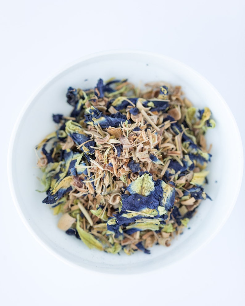 May include: A white bowl filled with a blend of dried herbs and flowers, including blue butterfly pea flowers, lemongrass, and other botanicals.