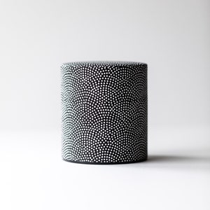 May include: A cylindrical container with a black base and a black body covered in a pattern of white dots. The container is set against a plain white background. The design is simple and modern.
