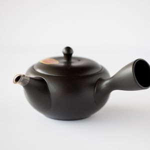 May include: A black ceramic teapot with a long spout and a lid. The teapot has a small, round body and a slightly curved spout.