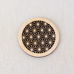 May include: A round wooden coaster with a geometric pattern cut out of the center. The pattern is a repeating design of stars and triangles.