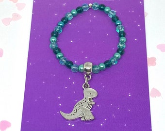 May include: A beaded bracelet featuring alternating translucent light blue and solid dark teal beads, centered with a silver-toned dinosaur charm. The bracelet is displayed on a purple card printed with the text JsJewelsHandmade JEWELLERY HANDMADE.