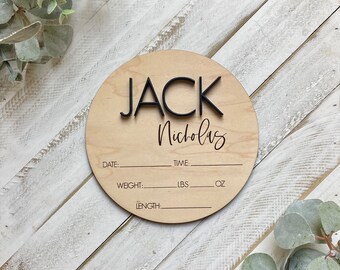 Puede incluir: Una placa redonda de madera con el nombre "JACK Nicholas" escrito en letras negras. La placa tiene espacios para rellenar la fecha de nacimiento del bebé, la hora, el peso en libras y onzas, y la longitud.
