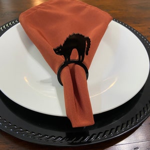 K&ouml;nnte beinhalten: Serviettenring aus Acryl in Form einer schwarzen Katze. Der Ring h&auml;lt eine rostfarbene Serviette. Der Ring befindet sich auf einem wei&szlig;en Teller mit einem schwarzen Untersetzer.