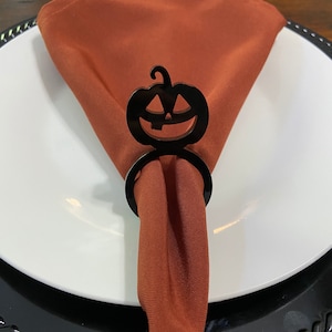 K&ouml;nnte beinhalten: Schwarzer Serviettenring in Form eines K&uuml;rbisses mit einem geschnitzten Jack-o'-Lantern-Gesicht. Der Ring h&auml;lt eine braune Serviette auf einem wei&szlig;en Teller mit einem schwarzen Untersetzer.