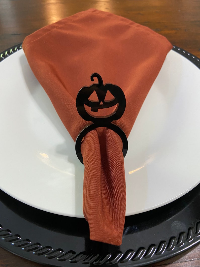 K&ouml;nnte beinhalten: Schwarzer Serviettenring in Form eines K&uuml;rbisses mit einem geschnitzten Jack-o'-Lantern-Gesicht. Der Ring h&auml;lt eine braune Serviette auf einem wei&szlig;en Teller mit einem schwarzen Untersetzer.