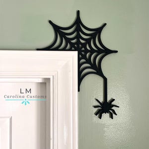 May include: Black spider web and spider corner decoration. The web is attached to a white door frame and a light green wall. The spider hangs below the web. The text "LM Carolina Customs" is on the door.