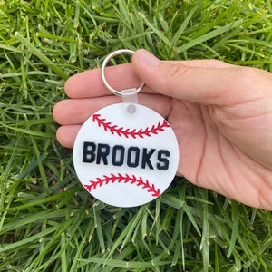 May include: White baseball-shaped keychain with red stitching and the name "BROOKS" in black lettering.