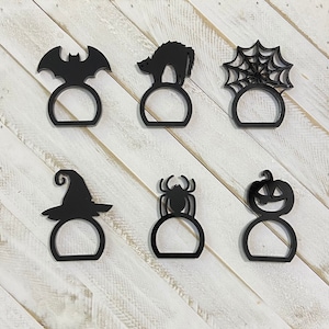 K&ouml;nnte beinhalten: Satz mit sechs schwarzen Serviettenringen zum Thema Halloween. Die Ringe zeigen eine Fledermaus, eine Katze, eine Spinne, einen K&uuml;rbis, einen Hexenhut und ein Spinnennetz.