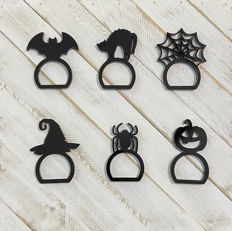 K&ouml;nnte beinhalten: Satz mit sechs schwarzen Serviettenringen zum Thema Halloween. Die Ringe zeigen eine Fledermaus, eine Katze, eine Spinne, einen K&uuml;rbis, einen Hexenhut und ein Spinnennetz.