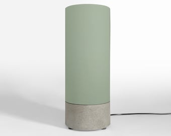 Może przedstawiać: Nowoczesna cylindryczna lampa stołowa z szałwiowym zielonym kloszem i betonową podstawą. Lampa jest podłączona i stoi na białej powierzchni. Lampa ma około 30 cm wysokości.