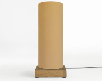 May include: A cylindrical table lamp with a tan lampshade and a square wooden base. The lamp is connected to a power cord. The design is minimalist and modern, suitable for a bedroom or living room.