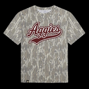 May include: A short-sleeved t-shirt with a camouflage pattern in shades of grey and tan. The word "Aggies" is displayed across the chest in a maroon script font with a white outline. The shirt has a crew neck and a small logo near the hem.