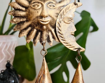 May include: A gold-colored metal wind chime featuring a sun and moon design. The sun has a smiling face, and the moon has a profile of a face. Two bell-shaped chimes hang below. The wind chime is suspended by a natural fiber rope.