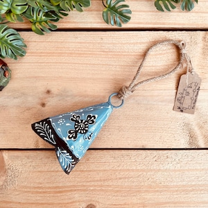 May include: A triangular metal bell painted in light blue with black and white floral patterns. The bell features a jute rope loop for hanging and a small rectangular brown paper tag with the text The Happy Hippie Bus printed on it.