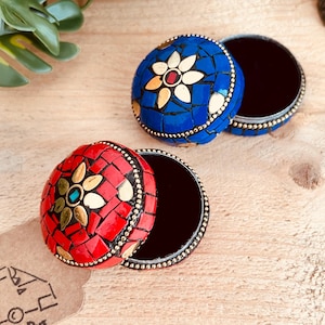 May include: Two round trinket boxes with mosaic lids, one in vibrant red and one in deep blue. Each lid features a central floral pattern made of gold-toned metal petals and a small centre stone, surrounded by small, irregular mosaic tiles.