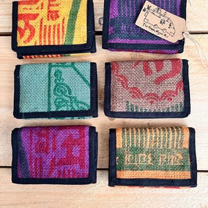 Puede incluir: Seis pequeñas carteras rectangulares hechas de un material tejido y texturizado. Las carteras presentan varios colores y patrones, incluyendo amarillo, morado, verde y marrón. Cada cartera tiene un borde negro y un cierre plegable. Una cartera tiene una etiqueta que dice "The Happy Hippie Bus."