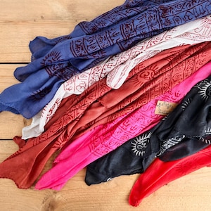 Könnte beinhalten: Eine Auswahl an farbenfrohen Schals in Blau, Weiß, Rot, Pink und Schwarz, jeweils mit einem sich wiederholenden Sonnen- und geometrischen Muster. Die Schals sind auf einer Holzoberfläche drapiert, wodurch ihr fließender Stoff zur Geltung kommt.