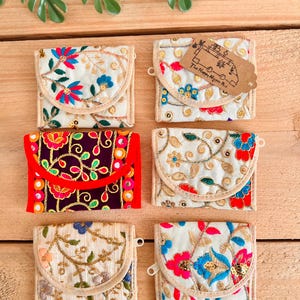 May include: Six small, embroidered fabric wallets with floral designs in various colours, including red, blue, and gold. The wallets have a flap closure and are arranged on a wooden surface. One wallet has a tag that reads "The Happy Hippie Bus".