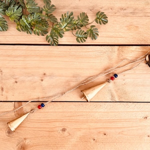 Puede incluir: Una guirnalda decorativa que presenta cuatro campanas cónicas de color latón suspendidas de un cordel de yute natural. Cada campana está acentuada con un par de pequeñas cuentas rojas y azules colocadas en el cordel, creando un diseño sencillo.