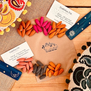 Puede incluir: Una variedad de conos de incienso en tonos naranja, rosa y gris, dispuestos sobre una superficie de madera. Los conos están acompañados de dos porta inciensos azules con diseños de estrellas y sol, y una bolsa de papel marrón con texto. También hay piezas de tela decorativas.